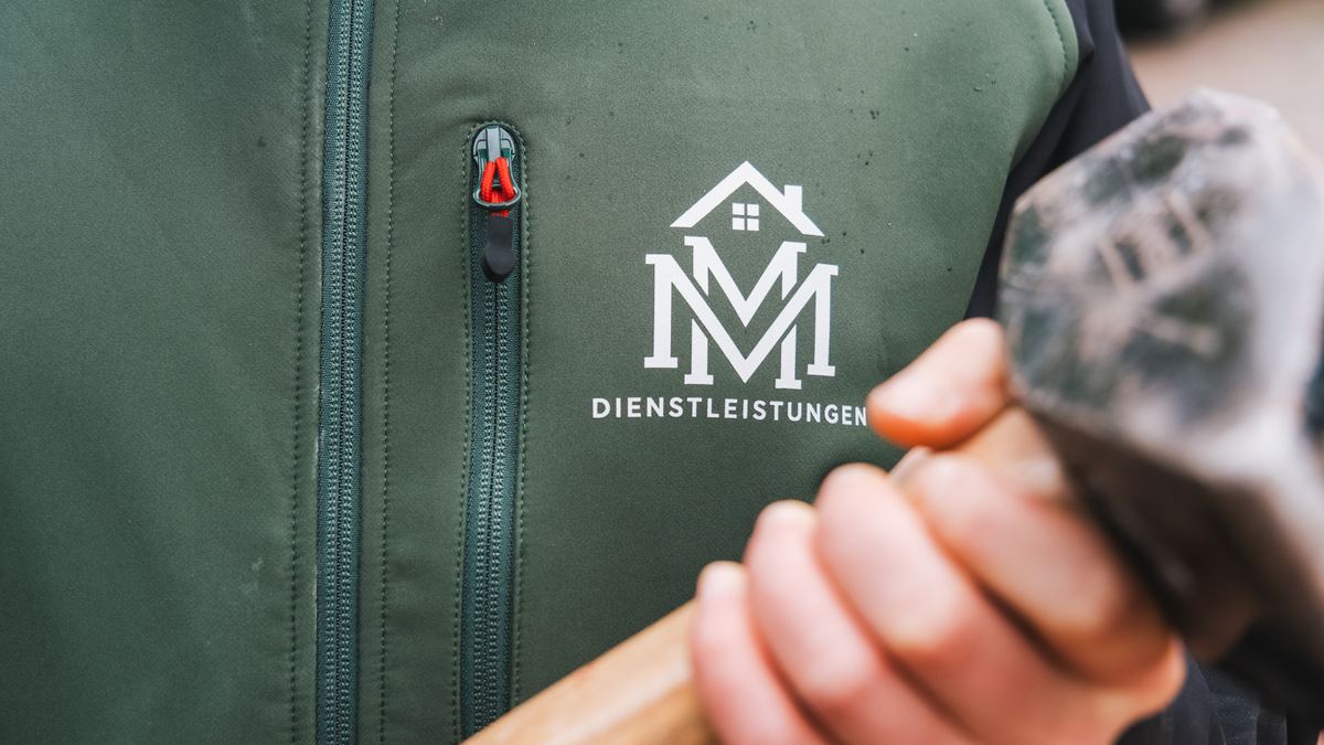 Hand eines MM-Dienstleistungen-Mitarbeiters mit Vorschlaghammer, im Hintergrund das Logo auf der Arbeitsjacke
