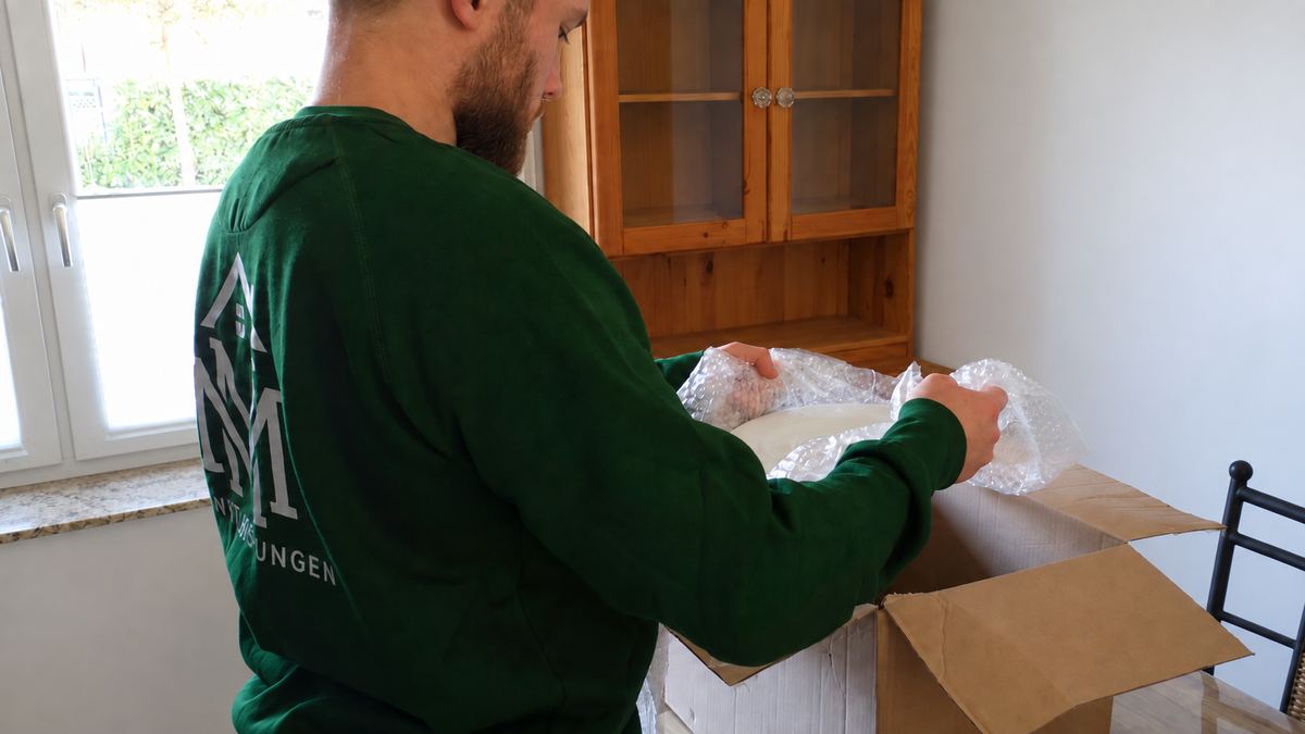 Ein Teammitglied von MM Dienstleistungen verpackt einen Gegenstand in Luftpolsterfolie über einem offenen Umzugskarton
